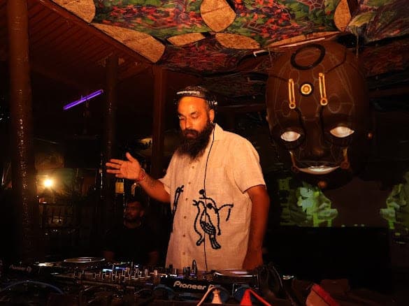 Immortal Baba behind Pioneer CDJs under a hand-painted psychedelic canopy, gesturing towards the floor, a large carved mask with lit eyes hanging to his right