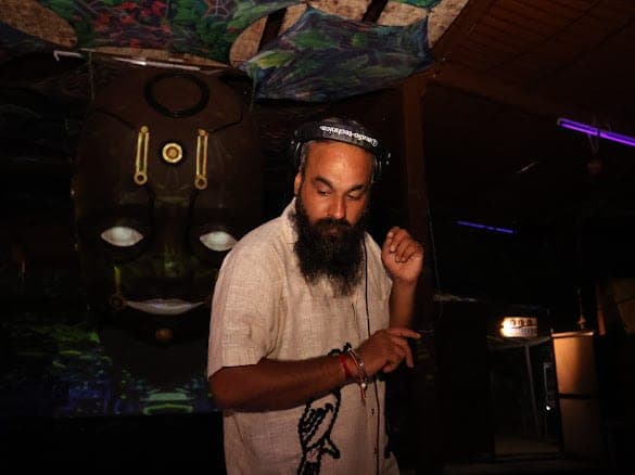 Immortal Baba standing at the decks, headphones on, beside a large carved wooden mask with glowing eyes, a violet laser crossing the canopy above